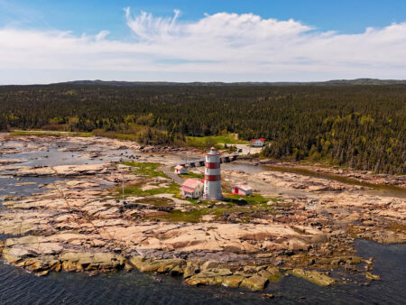 Vue aérienne du phare de Pointe-des-Monts, rouge et blanc, sur une pointe rocheuse dans le golfe du Saint-Laurent.