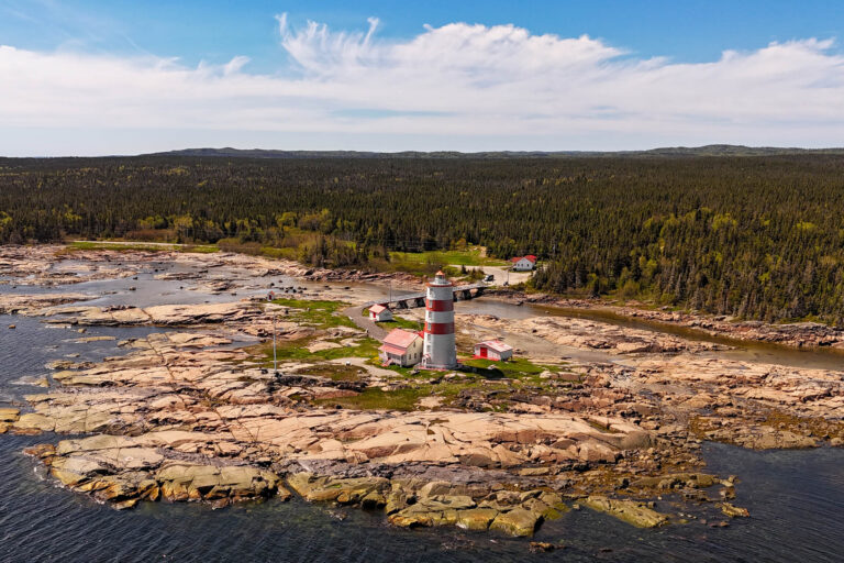 Vue aérienne du phare de Pointe-des-Monts, rouge et blanc, sur une pointe rocheuse dans le golfe du Saint-Laurent.