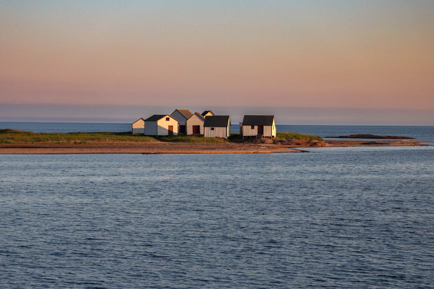 Hangars de pêche historiques blancs avec portes rouges sur une pointe rocheuse à Natashquan, Québec, au crépuscule.