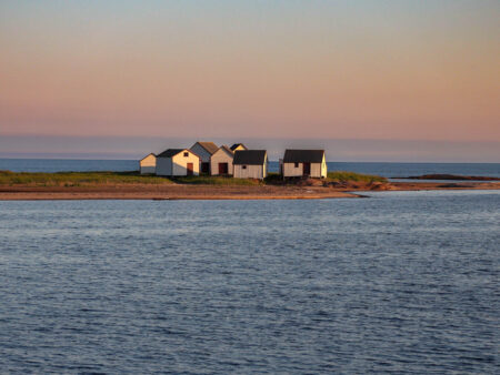 Hangars de pêche historiques blancs avec portes rouges sur une pointe rocheuse à Natashquan, Québec, au crépuscule.