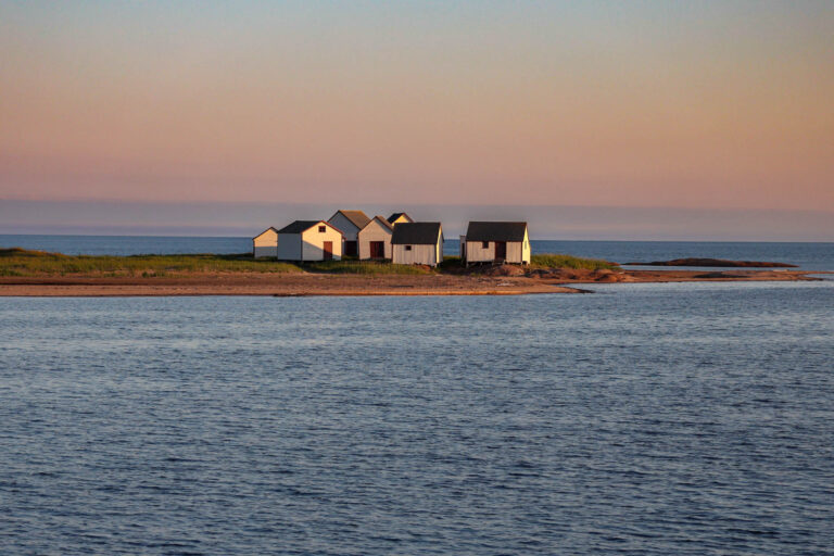 Hangars de pêche historiques blancs avec portes rouges sur une pointe rocheuse à Natashquan, Québec, au crépuscule.